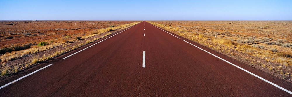 Stuart Highway stretches straight through an arid desert landscape with dry vegetation under a clear blue sky.