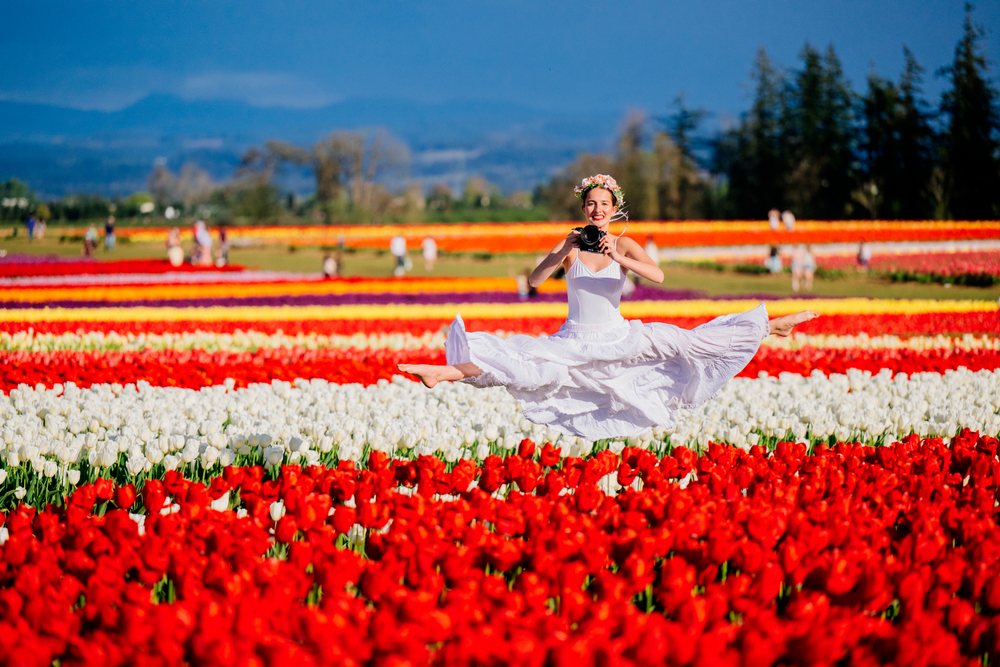 Ballet dancer wearing a flower crown leaps in a vibrant tulip field holding a camera, USA.