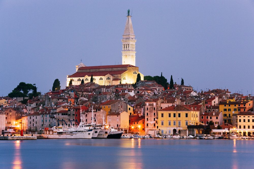 Dawn illuminates Rovinj town with a lit church bell tower by the harbor and boats along the waterfront, Croatia.