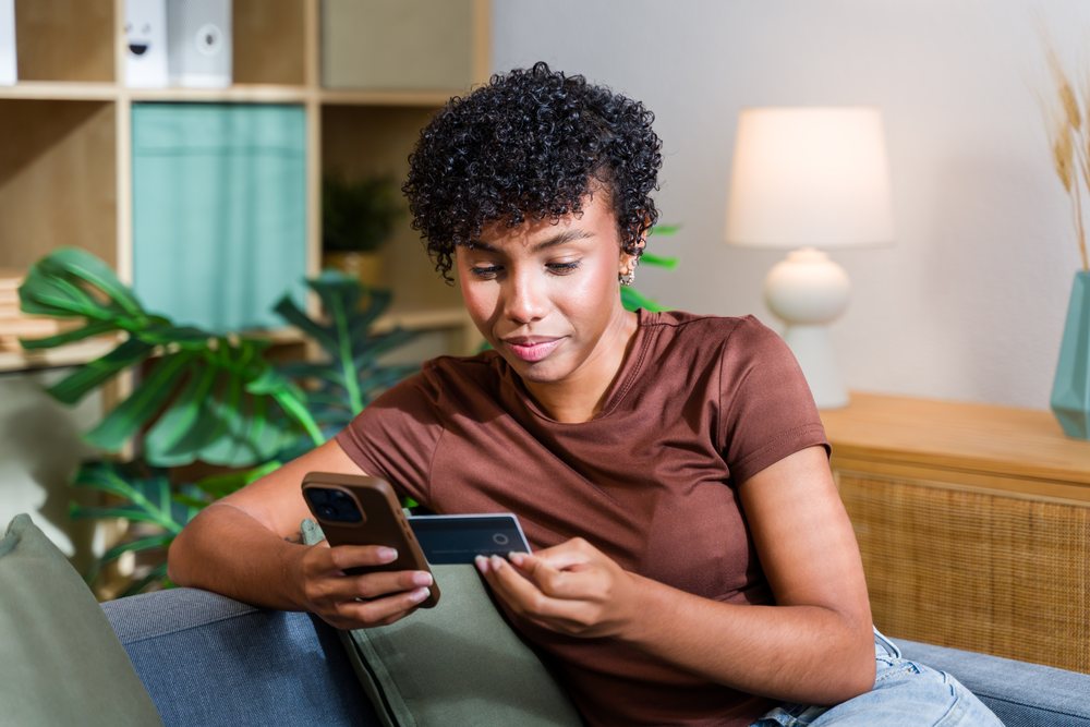 Woman sitting on a sofa at home using a smartphone and holding a credit card for online payment, Spain.