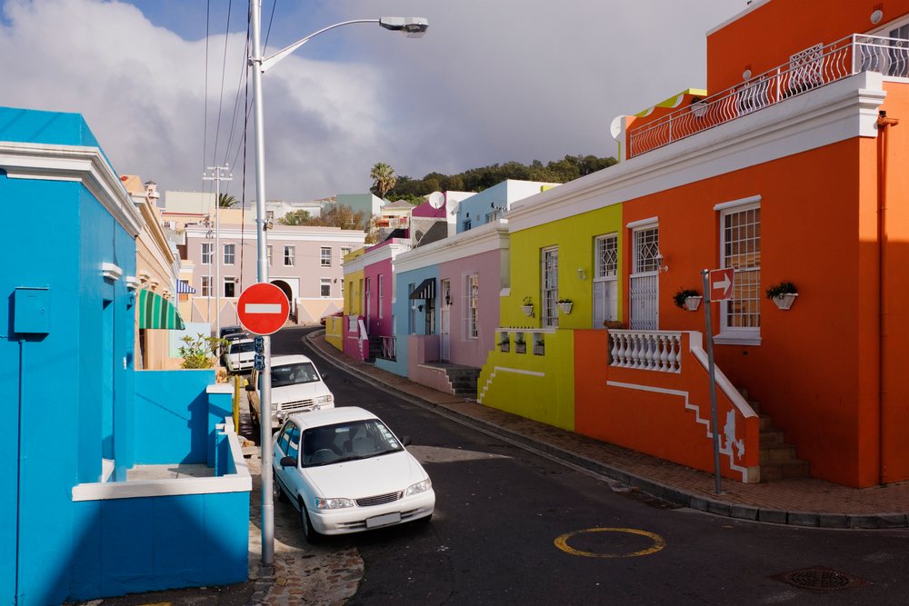 Colorful houses with vibrant painted walls line a curved uphill street in Bo Kaap, under a cloudy sky, Western Cape, South Africa.