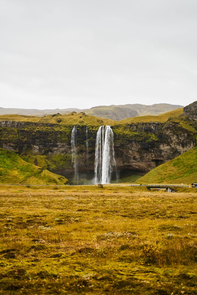 A wide waterfall cascades down rocky cliffs into a misty valley surrounded by grassy hills, Iceland.