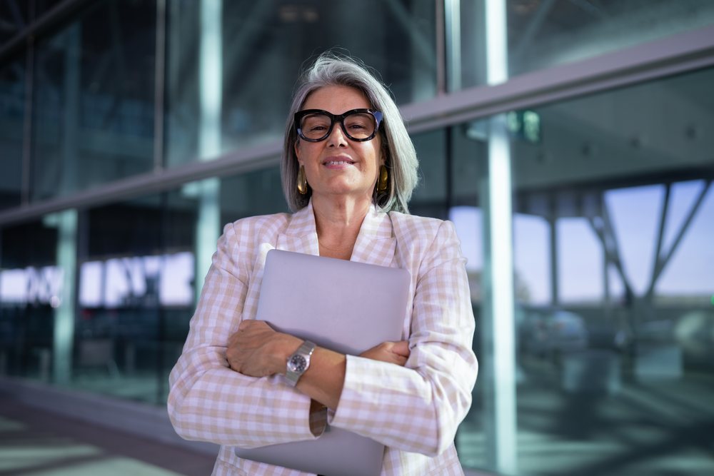 Businesswoman with gray hair and glasses confidently holding a laptop outside a modern glass building in an urban setting, Spain, Madrid.