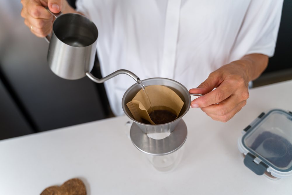 Person pouring hot water from a metal kettle over coffee grounds in a metal dripper with a filter on a kitchen counter, Spain.