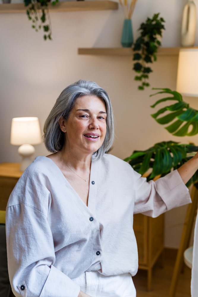Gray-haired woman smiles indoors near a monstera plant and lamp in a cozy living room with wooden furniture, Spain.