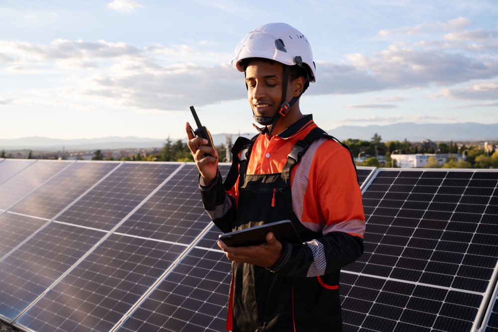 Worker wearing helmet and safety gear inspects solar panels outdoors in Madrid using walkie talkie and tablet at sunset, Spain.