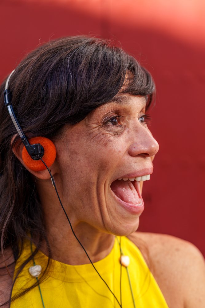 Woman wearing orange headphones speaking or singing with a joyful expression against a red background, retro, vintage, Málaga, Spain.