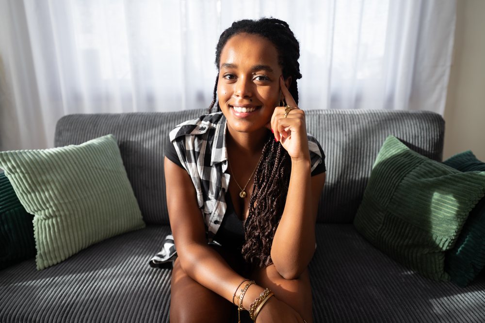 Smiling woman with long braids sits on a gray couch with green pillows in a cozy indoor living room setting, moving, Spain.