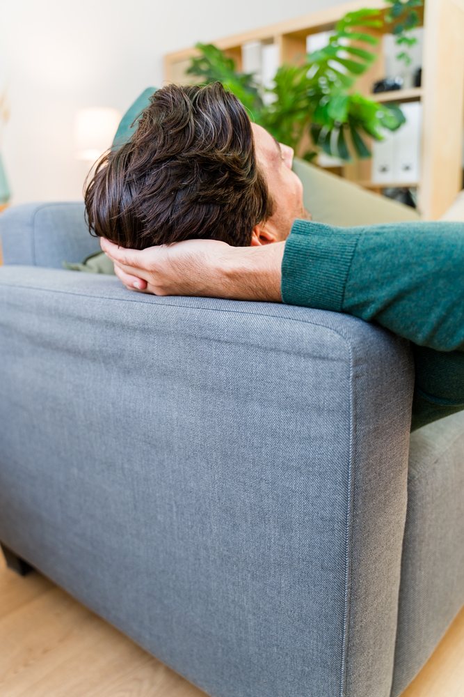 Man wearing a green sweater relaxes on a grey sofa in a modern living room with wooden flooring and houseplant, Spain.
