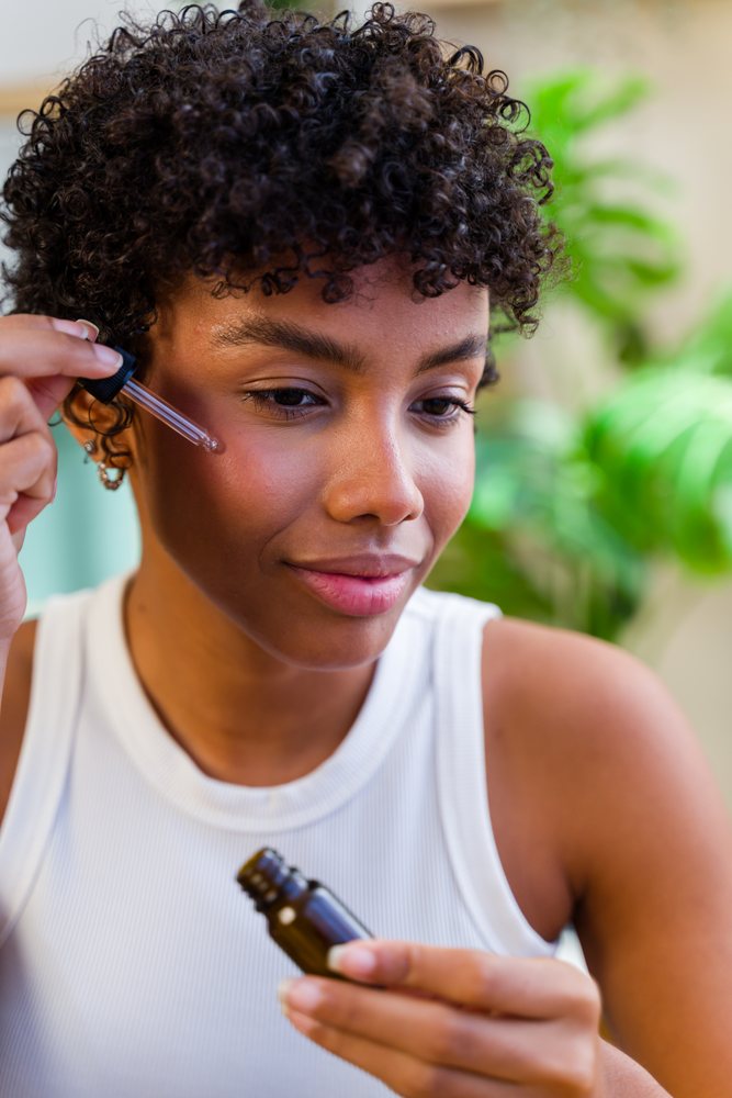 Woman indoors applying face serum drops to her cheek with green plants in the background, Spain.