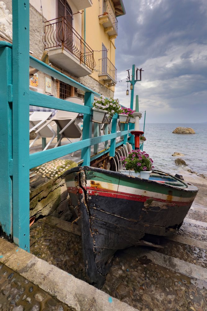 Old Fishing Boat on stone steps along a Narrow Street with flower pots by the Mediterranean Sea under a stormy sky, Scilla, Calabria, Italy.