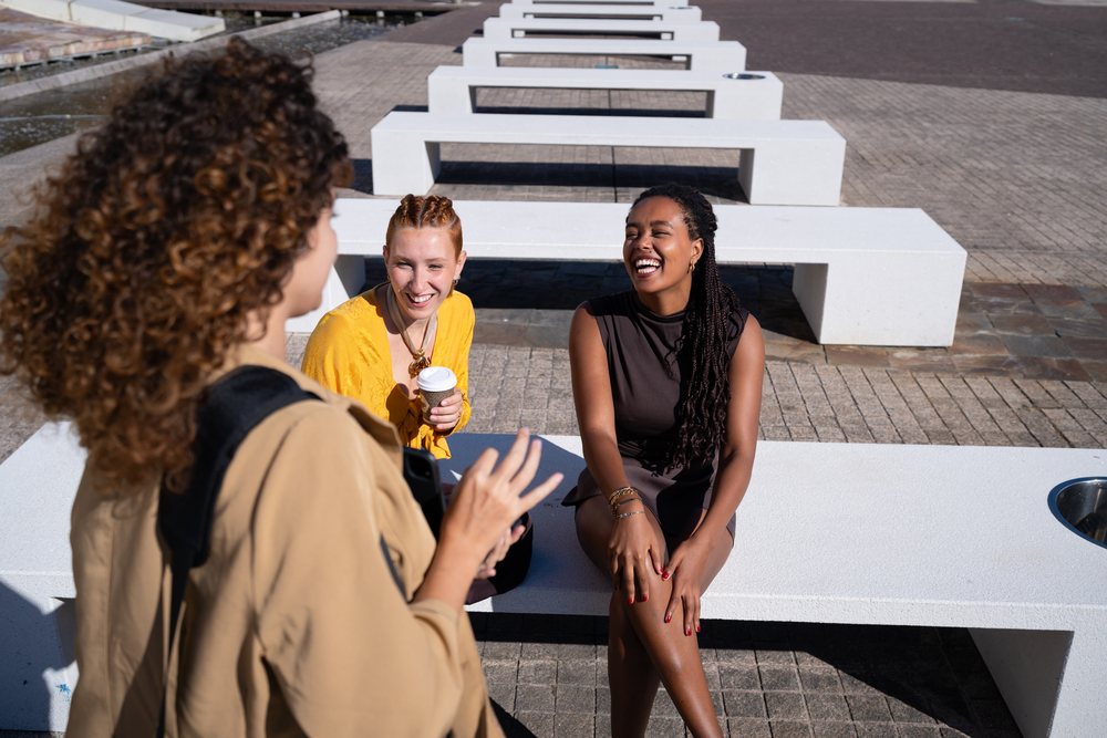 Three women sitting on white benches, laughing and chatting in an urban outdoor plaza under daytime sunlight, Spain.