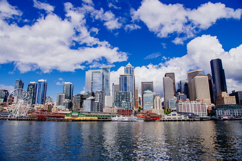 City skyline featuring the Seattle waterfront, ferris wheel, boats, piers, and reflections under a blue sky with clouds, USA.