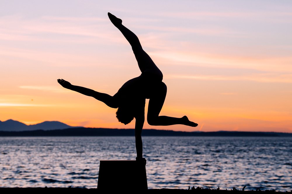 Person performs a one-handed handstand silhouette on a raised platform by the ocean at sunset with mountains in the background, USA.