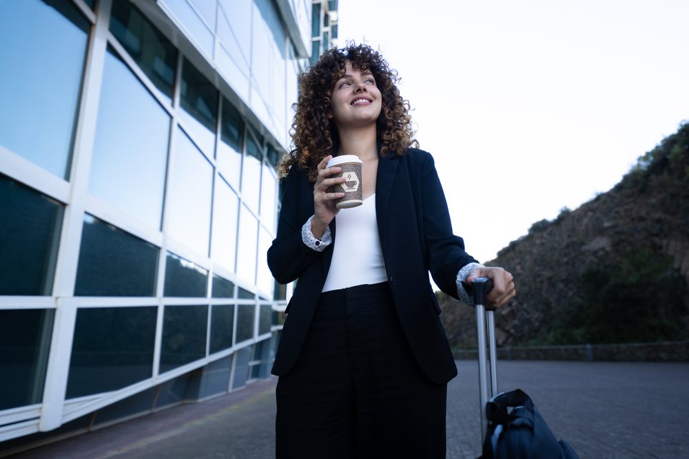 Businesswoman holding a coffee cup and pulling a suitcase outside a modern glass building during a business trip, Spain, Santiago De Compostela.
