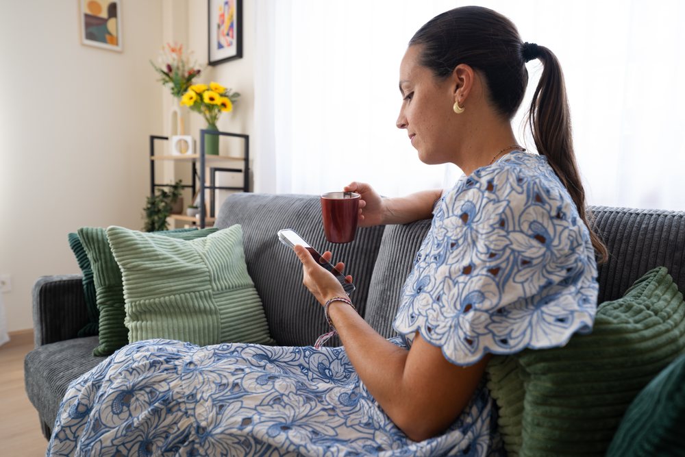Woman wearing a blue and white floral dress sits on a sofa holding a mug and using a smartphone in a bright living room, telecommuting, Spain.