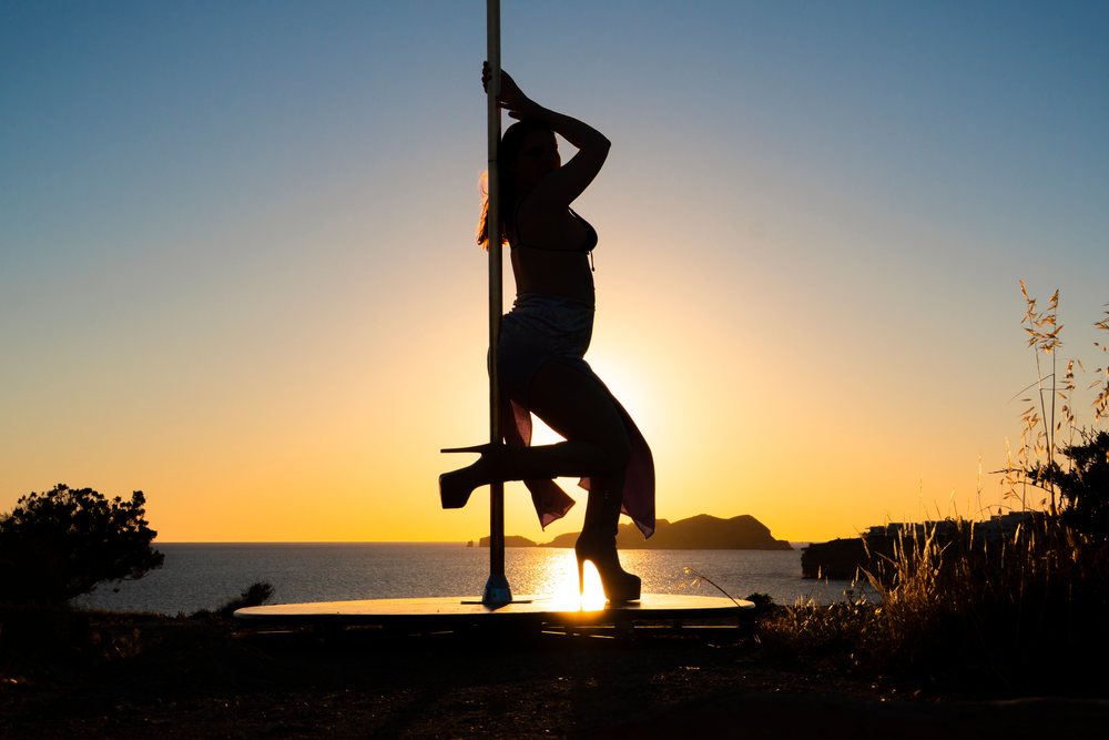 Silhouetted pole dancer in high heels and skirt performs on an outdoor platform at sunset by the calm sea shore of, pole dance, empowerment, Ibiza, Spain.