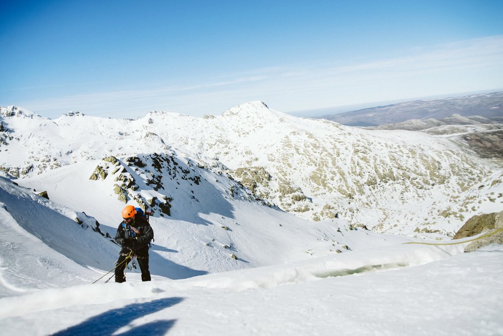 Mountaineer climbing a snowy mountain slope with rope and helmet in Spain's mountainous landscape under a clear blue sky.
