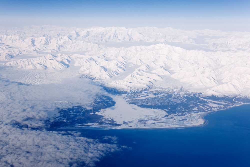 Aerial view showing snow-covered Alaskan coastline with glaciers, mountains, ocean, and clouds.