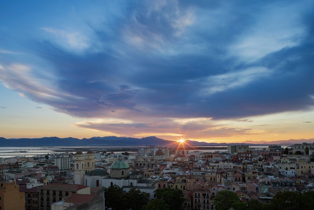 Sunrise behind mountain range with city skyline of Old Town, showing buildings with domes and roofs near the sea, Travel Photography, Cagliari, Sardinia, Italy.
