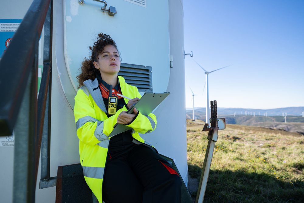 Woman technician wearing high visibility jacket holding clipboard and walkie talkie sitting beside wind turbine at a wind farm in hilly, sustainability, renewable energy, Galicia, Spain.
