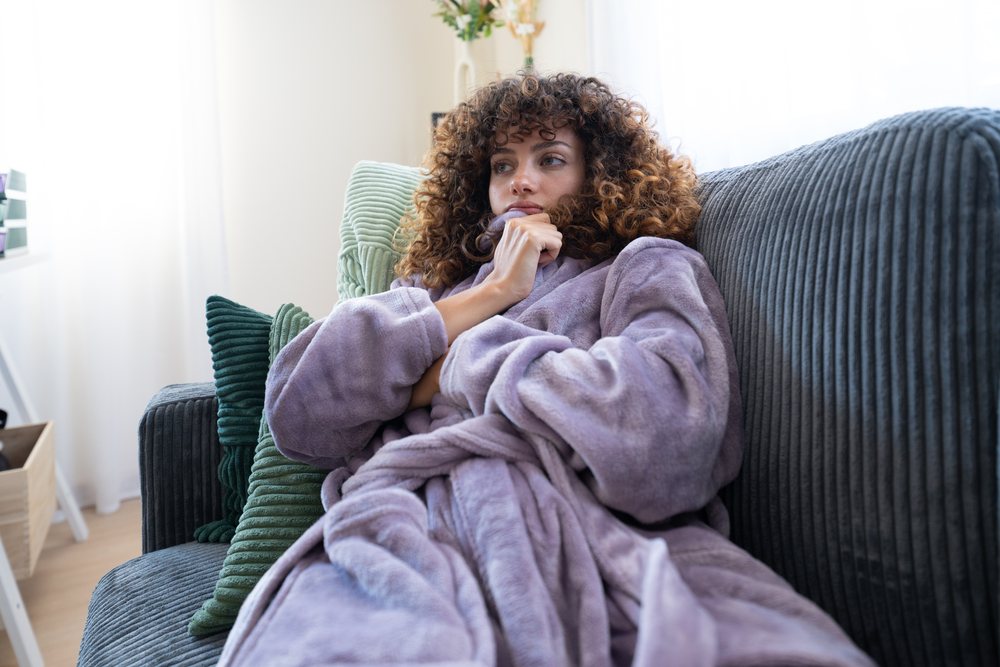 Woman sitting on a couch indoors at home, wearing a purple robe and resting thoughtfully in natural daylight, Spain.