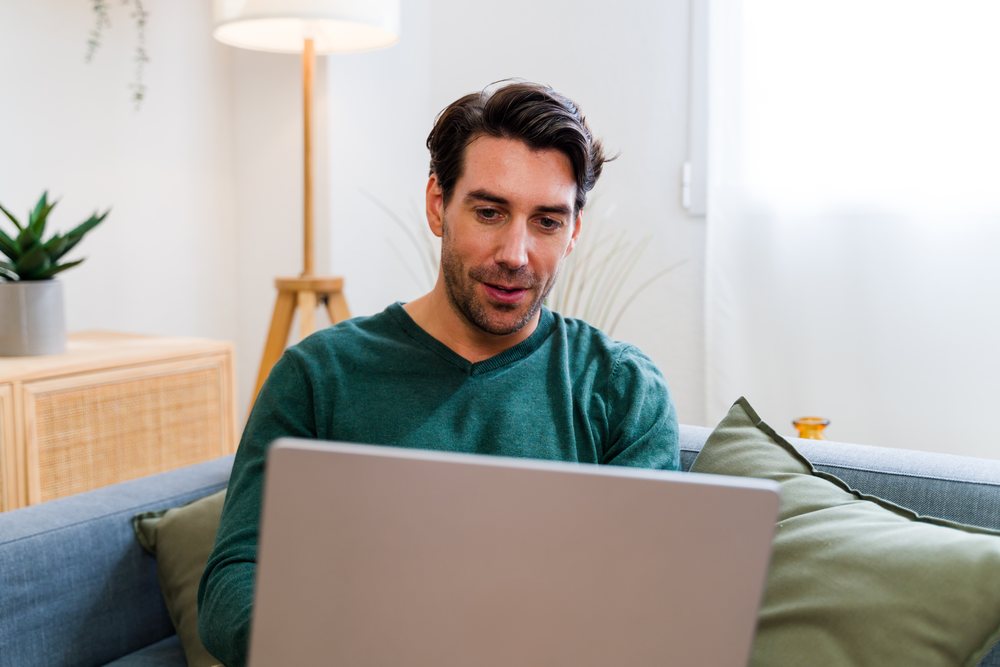 Man wearing green sweater using a laptop while seated on a couch in a bright living room with wooden furniture and indoor plants, Spain.