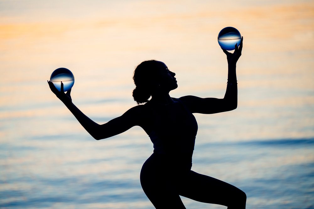 Silhouette of a woman holding and balancing two crystal balls at sunset over water, yoga, acrobatics, USA.