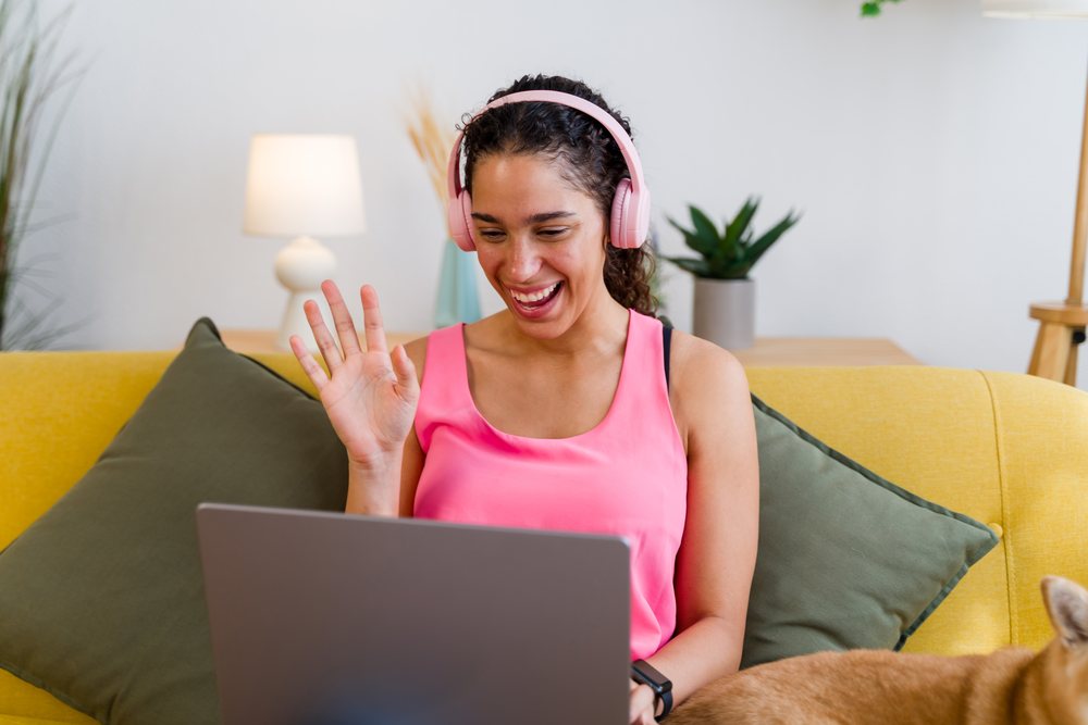 A woman wearing pink headphones waves and smiles during a video call on her laptop while sitting on a yellow sofa in a cozy living room, Spain.
