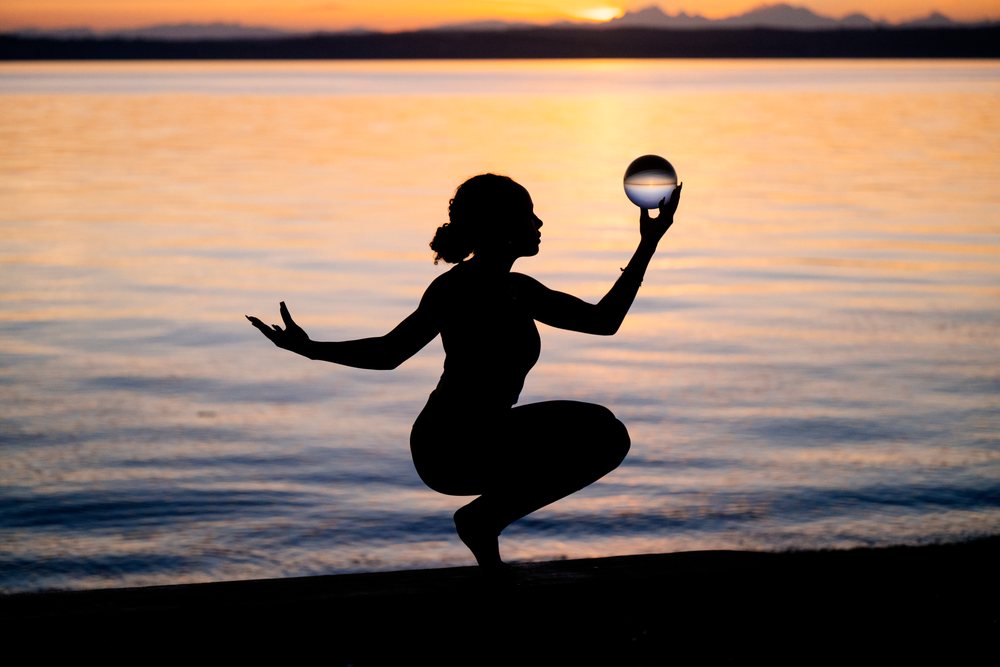 Silhouette of a woman performing a yoga balance pose while holding a glass sphere at sunset on a lake shore, USA.