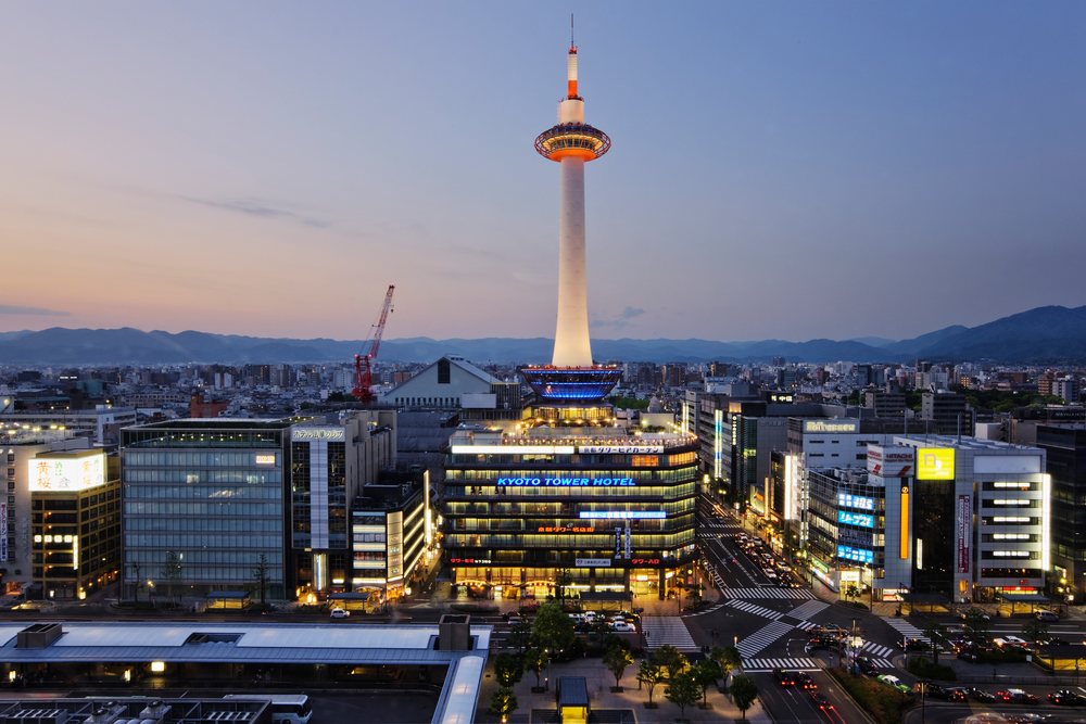 Kyoto Tower illuminated above the cityscape with buildings, streets, cars, and mountains visible at evening, Honshu, Japan.