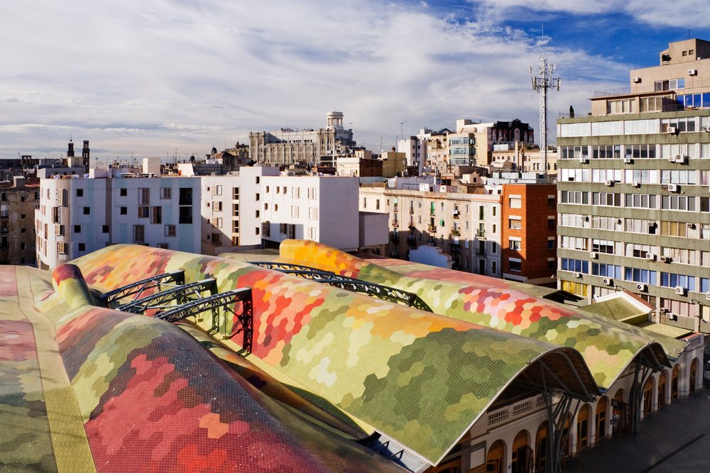 Santa Caterina Market's colorful mosaic roof stands out amid Barcelona's urban buildings in bright daylight, Spain.