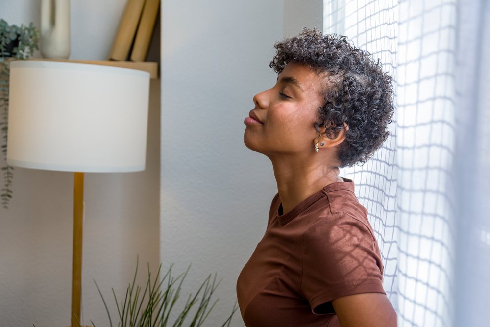 Young woman with curly hair wearing a brown shirt relaxes indoors near a window with natural light and calming decor.