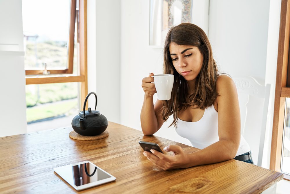 A white woman drinks tea while using her smartphone indoors