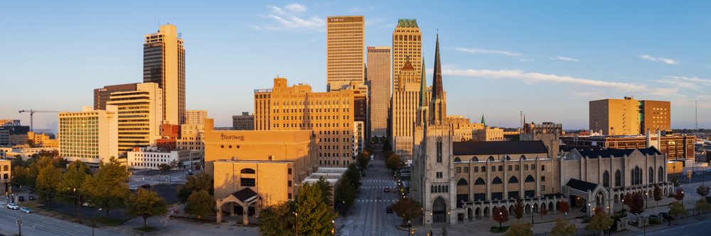 Early morning sunrise illuminates Tulsa skyline with tall buildings, church spires, streets, and trees along historic Route 66, Oklahoma, USA.