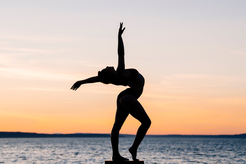 Silhouette of a female dancer posing with outstretched arms on a platform by the ocean at sunset, USA.