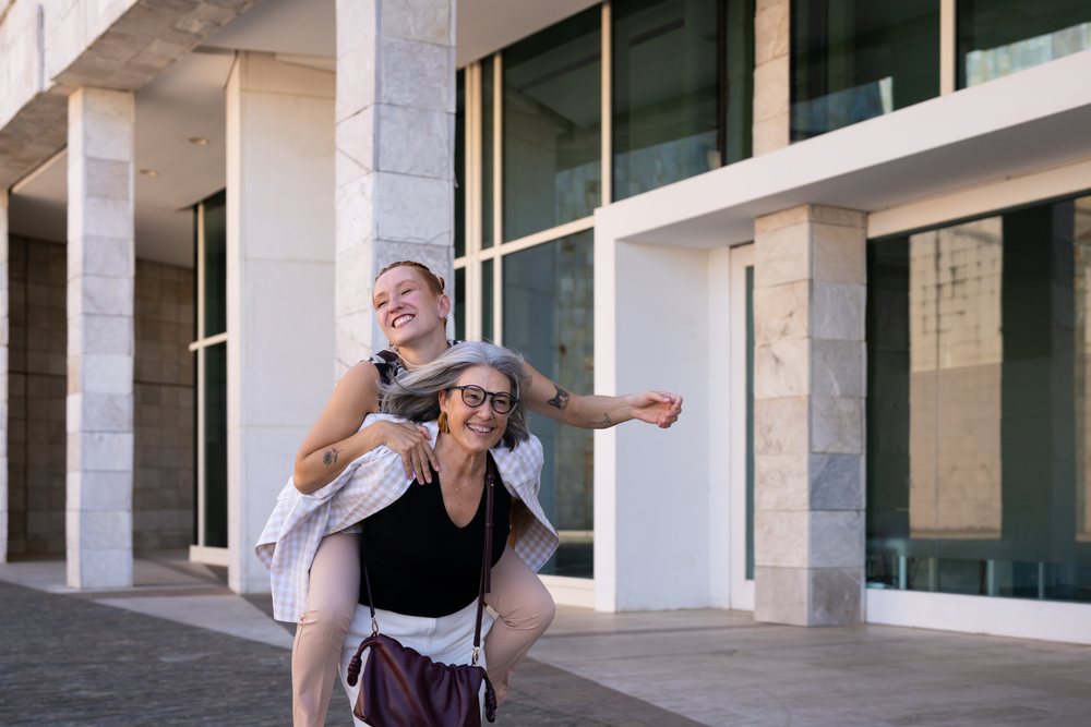 Two women share a joyful piggyback moment outside a modern building, businesswomen, Spain.