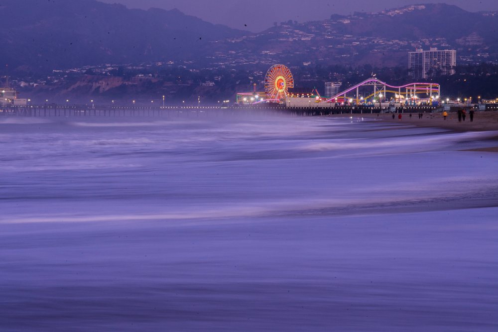 Santa Monica Pier illuminated at dusk with ocean waves washing onto the nearby sandy beach and mountains visible in the background, CA, USA.