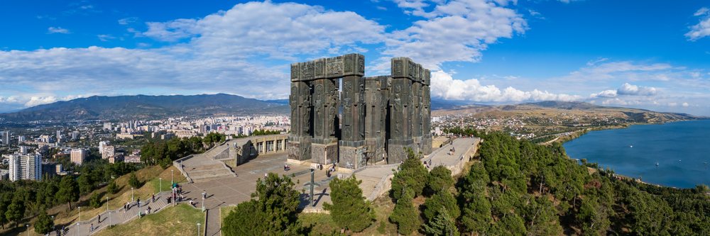 Historic Georgia monument with tall sculpture columns on Keeni Hill overlooks Tbilisi cityscape and a nearby lake under a partly cloudy sky, Cityscapes.