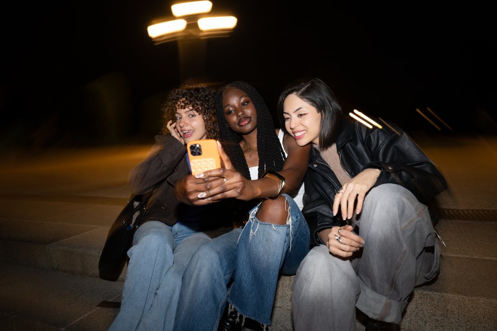 Three women sitting on outdoor steps in Madrid taking a selfie at night under street lighting, Spain.