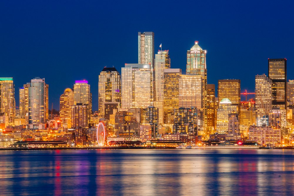 Nighttime Seattle skyline with illuminated skyscrapers and a ferris wheel reflecting on the water under a clear sky, USA.