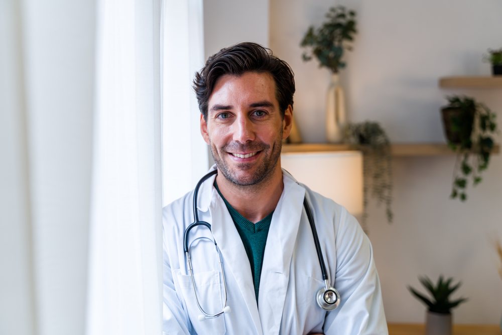 Male doctor standing and smiling indoors in a medical office wearing a white coat and stethoscope, Spain.