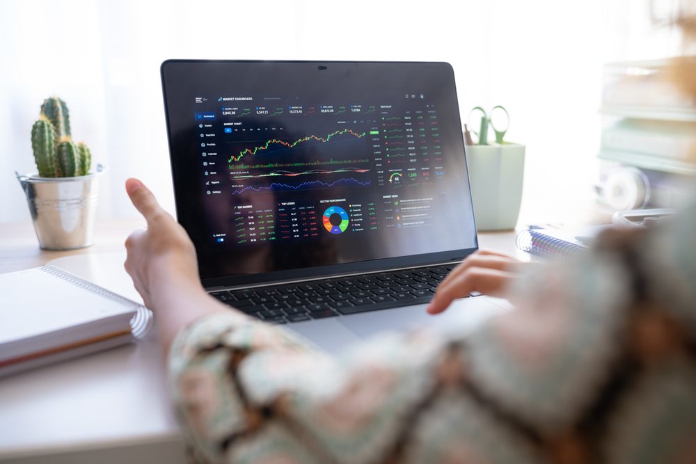 Woman points at a stock market dashboard displayed on a laptop in a home office with a notebook and cactus plant on the desk, Spain.