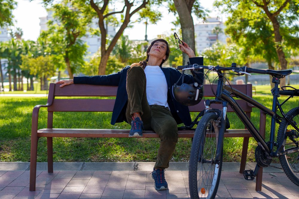 Woman sitting on a wooden bench holding eyeglasses beside her bicycle with helmet hanging on handlebar in a sunny park, Málaga.