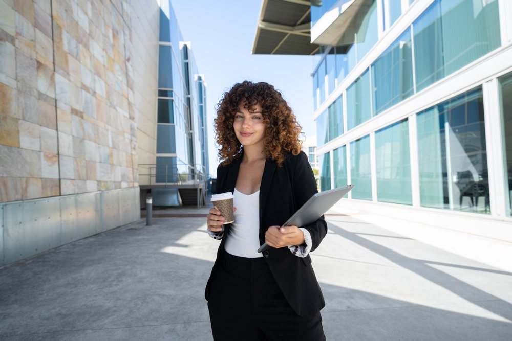 Businesswoman holding coffee cup and laptop walking outside a modern office building, traveling executive, Spain.