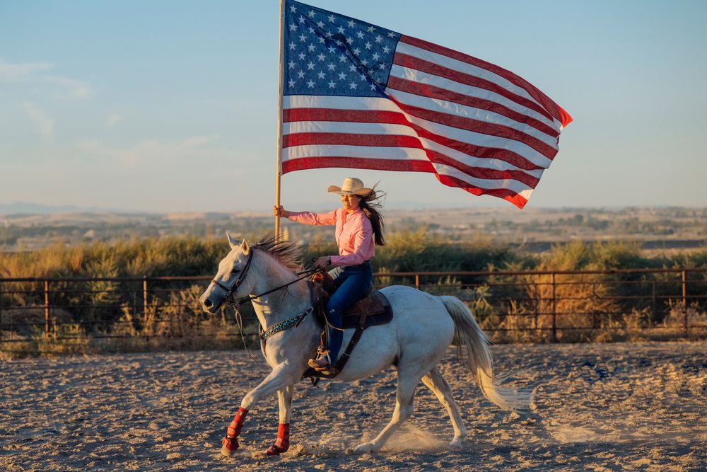 Woman wearing a cowboy hat rides a white horse while holding an American flag in a sandy Southwest USA desert at sunset.