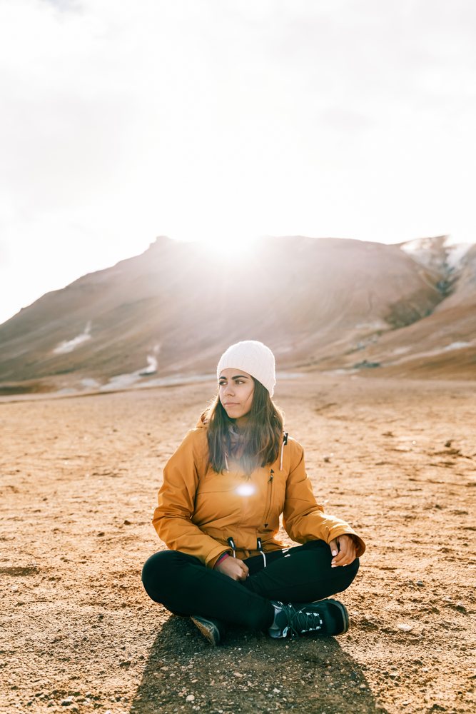A woman wearing a yellow jacket and white knit hat sits cross-legged on rocky ground with mountains and bright sunlight in the background, Iceland.