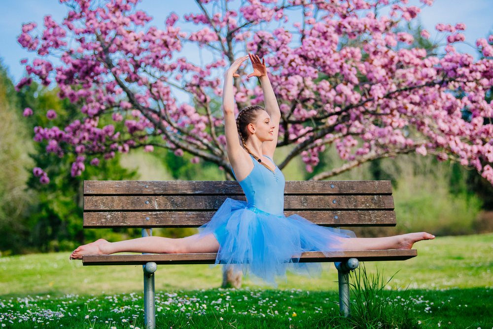 Female ballet dancer wearing a blue tutu performs a split on a wooden bench beneath blooming cherry blossoms in a grassy park, USA.