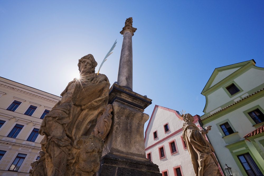Statues and a tall column stand in a historic Bohemian town square beneath a clear blue sky with sun flare, Bohemia, Czech Republic.