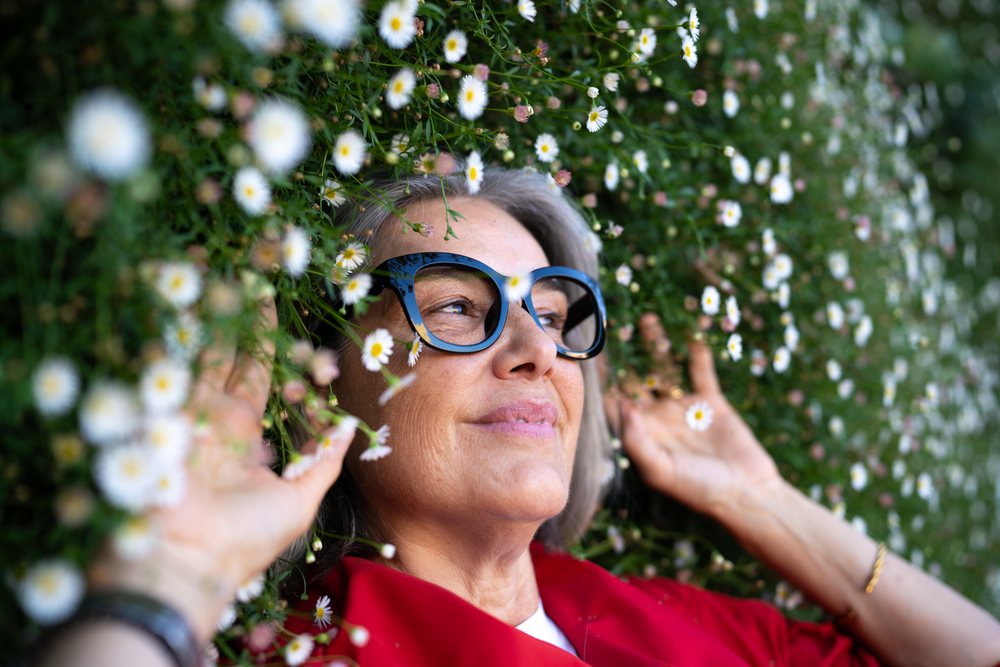 Mature woman with gray hair wearing glasses lies peacefully among white daisies and greenery outdoors, smiling gently, Spain.
